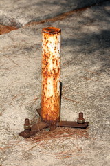 Short rusted metal pole mounted on concrete floor with strong screws and bolts used as foundation for parasol surrounded with dry fallen pine needles on warm sunny day