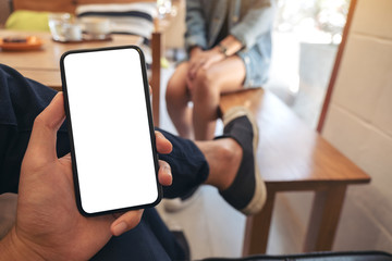Mockup image of a man's hand holding black mobile phone with blank white screen with woman sitting in cafe