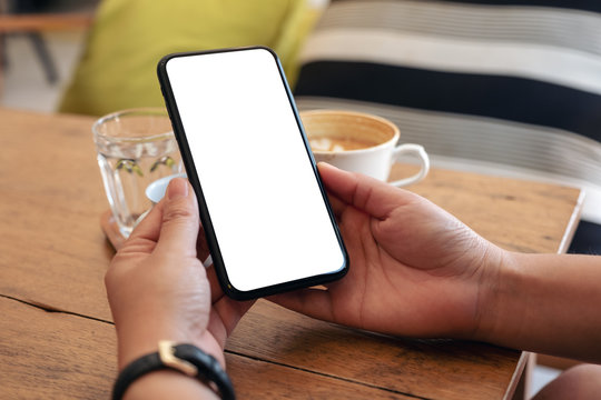 Mockup image of hands holding black mobile phone with blank screen with coffee cup on wooden table in cafe