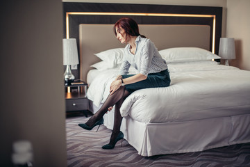Elegant red-haired lady touching with arms her legs, being tired from wearing stilettos while sitting in bedroom after returning home from office.