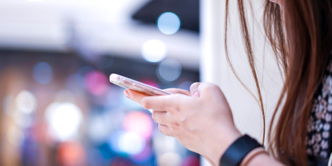 Young girl is holding smart phone in her hand (beside angle/ view); playing, typing, chatting, emailing with blurry bokeh department store behind.