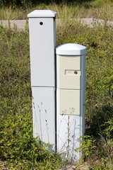 Two narrow tall light grey locked electrical boxes surrounded with tall uncut grass and other vegetation at abandoned industrial complex on warm sunny day