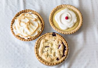 Assorted delicious whole Pies sitting on a table. A view of three different homemade pies - Lemon Meringue, cream and berry pies. - obrazy, fototapety, plakaty