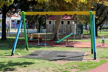 Wooden outdoor public playground equipment in shape of vintage looking swing with rubber protection on ground and other equipment in background surrounded with protective rubber and trees in backgroun