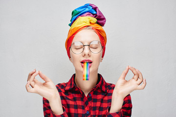 A pretty young woman with glasses and full lips in a turban and a red shirt holds a rainbow in her mouth and meditates against a white wall. LGBTQ Lesbian, gay, bisexual, transgender, queer.