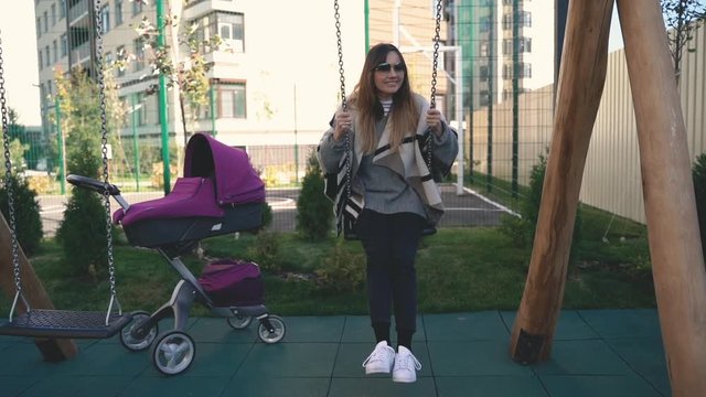 A middle plan of woman riding a swing in slowmo, a burgundy baby pram is near.