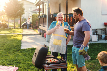 Cheerful father and son drinking beer while standing next to grill in backyard at summer. Family gathering concept. - obrazy, fototapety, plakaty