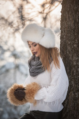 Attractive woman with white fur cap and jacket enjoying the winter. Side view of fashionable blonde girl posing against snow covered bridge. Beautiful young female with cold weather outfit