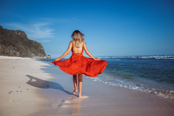 Girl walking on the beach with white sand in a red dress on the background of the ocean, the view from the back without a face.