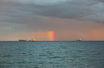 Short rainbow over sea, between three big tankers, under heavy clouds
