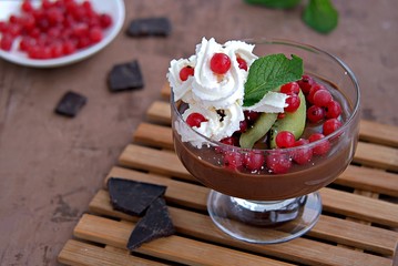Chocolate pudding with whipped cream, red currants and kiwi in a glass ice-cream bowl on a brown concrete background.
