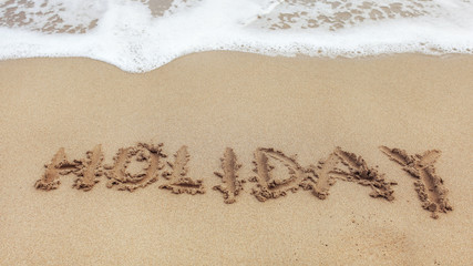 Word HOLIDAY written on wet sand of beach, small spot of white sea water in top part.