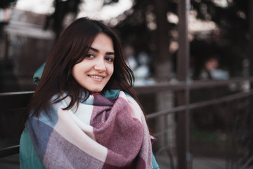 Beautiful young girl in winter clothes standing back to the fence near cafe on the street and looking far