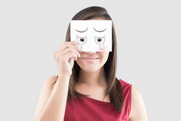 Asian woman holding a white paper with a cartoon cry face on it in front of her eyes against the gray background