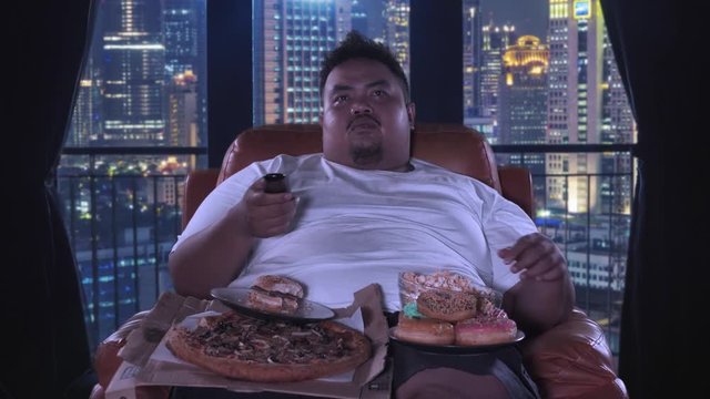 Greedy fat man watching TV while eating junk foods on the sofa in apartment. Shot in 4k resolution