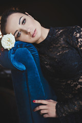Gorgeous woman in lace dress is holding rose and sits in armchair into expensive room