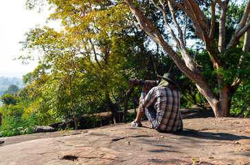 Copy space. Men wearing a hat are sitting shooting a camera on rocky cliff.