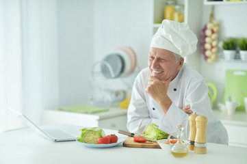 Portrait of elderly male chef cooking at table with laptop