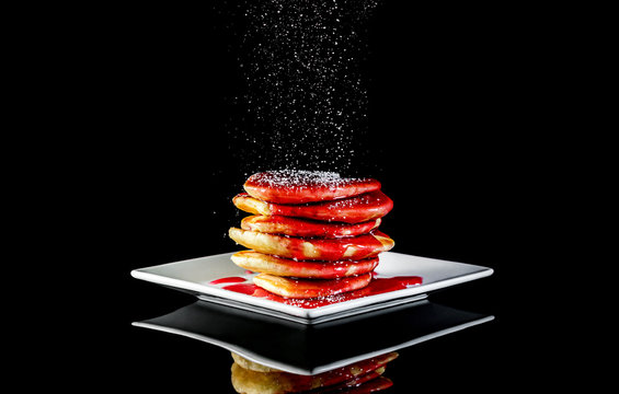 Powdered sugar sprinkles to stack of pancaces on black reflective background.