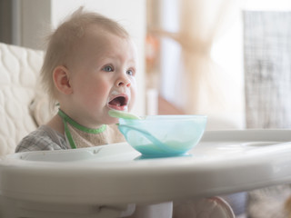 one year old boy learns to eat by himself. the kid eats porridge from a blue plate while sitting in a highchair, his face is soiled in porridge