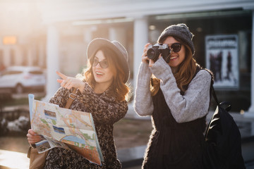 Stylish girls spending time outdoor in cold day exploring new places with camera. Gorgeous female photographer walking around city with sister which pointing with finger away and smiling holding map.
