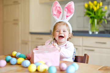 Cute little toddler girl wearing Easter bunny ears playing with colored pastel eggs. Happy baby child unpacking gifts. Adorable healthy smiling kid in pink clothes enjoying family holiday