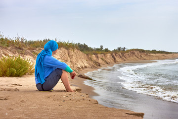 A woman in a blue scarf and a sweater on her head is sitting on the beach by the sea