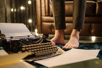 closeup photo of man's legs near a vintage typewriter on a piece of newspaper and dark blue velvet fabric on the floor in a dark room with light bulbs near a brown leather sofa