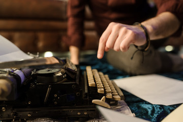 lateral view of man's hand typing on vintage typewriter on a piece of newspaper and dark blue velvet fabric on the floor in a dark room with light bulbs
