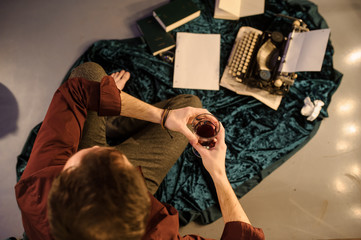 top view of man with a glass of wine in his hands sitting near a vintage typewriter on a piece of newspaper and dark blue velvet fabric on the floor with sheets of paper and books arround