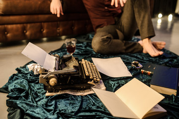 lateral view of man sitting near a typewriter on a dark blue velvet fabric on the floor in a dark room with light bulbs near a brown leather sofa with sheets of paper and books arround