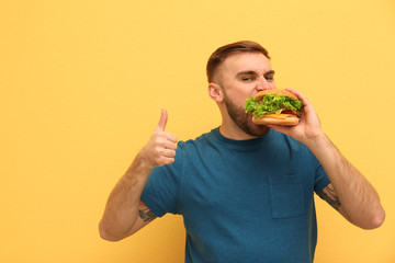 Young man eating tasty burger on color background. Space for text