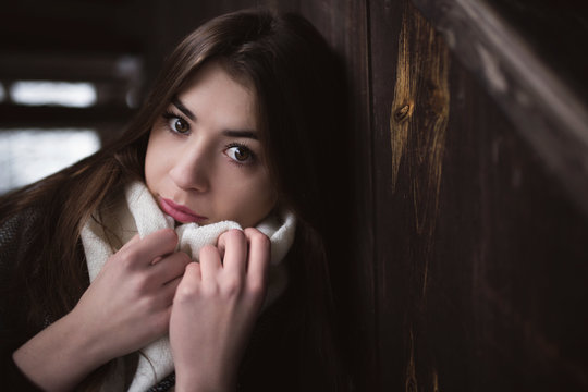 Portrait Of Pretty Girl With Beautiful Look Into Camera And Delicate Skin, She Keeps Hands Close To Her Face On Scarf. Cold Winter Season. Interior Of Abandoned House Or Shed With Dark Wooden Walls.