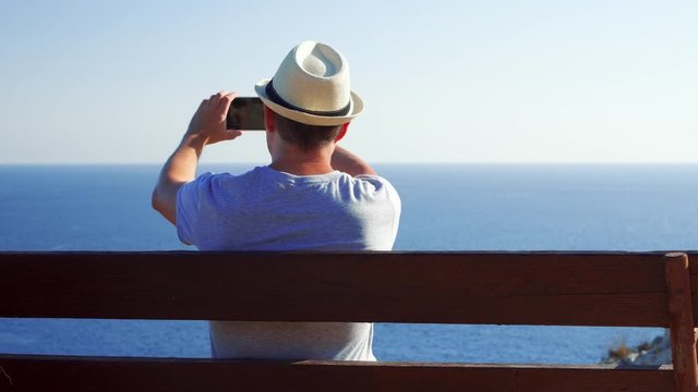 Young handsome man sitting relaxing on bench at edge of cliff take pictures on cellphone. Male in hat enjoying breathtaking view of blue Mediterranean sea making photos on mobile phone