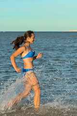 A sexy young brunette woman or girl wearing a bikini running through the surf on a deserted tropical beach with a blue sky. Young woman running by the sea. vertical photo