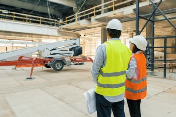 Building workers working at construction site, builders looking in blueprint, back view, copy space. Building, development, teamwork and people concept