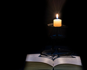 Opened book with eyeglasses, near are glowing candle, all things on dark background