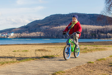 six-year-old boy in a jacket, scarf and hat rides a two-wheeled children's bike on the sidewalk on the background of the mountains and the sea