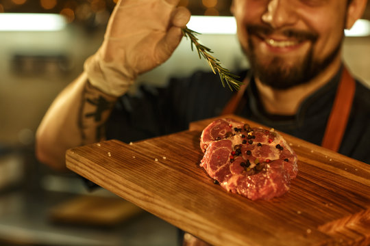 Tasty Beef Steak With  Salt And Pepper On Wooden Cutting Board. Chef Decorating Meat With Rosemary. Smiling Man With Bearded Face, In Gloves And Tattoo On His Arm On Background.