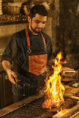 Chef concentrated on process of cooking delicious beef steak on grill. Steak flaming with fire and smoke. Beautiful bearded man wearing jeans apron on kitchen in restaurant.