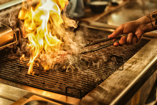 Front view of delicious juicy steak flaming with fire and smoke on grill. Hand of professional chef turning over steak. Concept of culinary and restaurant food and kitchen.
