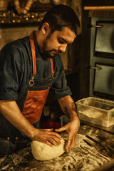 Hands of professional baker making dough for white homemade bread. Table in kitchen covered with flour. Man with bearded face wearing jeans apron.  Traditional cuisine.