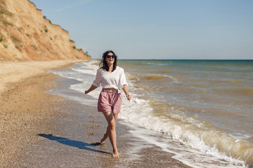 Happy young woman in white shirt and sunglasses running and having fun with waves on sunny beach. Hipster slim girl relaxing and smiling at sea. Summer vacation concept. Copy Space. Joyful moment