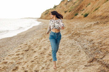 Happy young boho woman running and having fun on sunny beach with rock. Hipster slim girl relaxing and smiling at sea. Summer vacation concept. Copy Space. Joyful moment