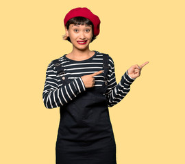 Young woman with beret frightened and pointing to the side over yellow background