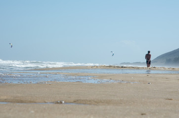 Close-up. Sea beach with the oncoming wave. Wet sand On the average plan is a man, he looks into the distance. In the background are the kitesurf sails. Sunny windy day.