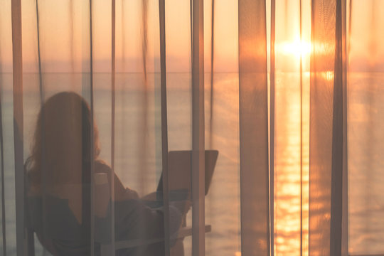 Naked beautiful female wrapped in a blanket sitting on a chair with laptop behind the transparent curtains on the glass balcony early morning at sunrise. Sea view