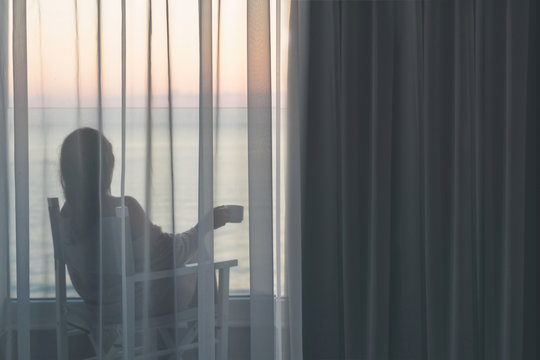 Naked beautiful female wrapped in a blanket sitting on a chair with cup of coffee behind the transparent curtains on the glass balcony early morning at sunrise. Space for text