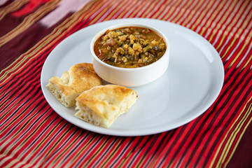 A bowl of hearty soup with chunks of vegetables and grains, served with pieces of crusty bread on a white plate, accompanied by a wooden utensil, set against a vibrant red and gold striped background.