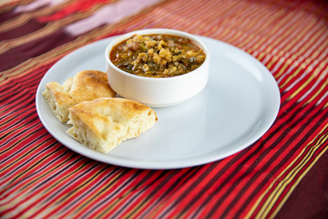 A bowl of hearty soup with chunks of vegetables and grains, served with pieces of crusty bread on a white plate, accompanied by a wooden utensil, set against a vibrant red and gold striped background.